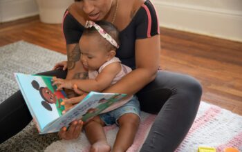 Eine Frau liest einem Baby ein Kinderbuch vor, während sie zusammen auf einem Teppich sitzen. Das Baby zeigt auf die bunten Bilder im Buch.