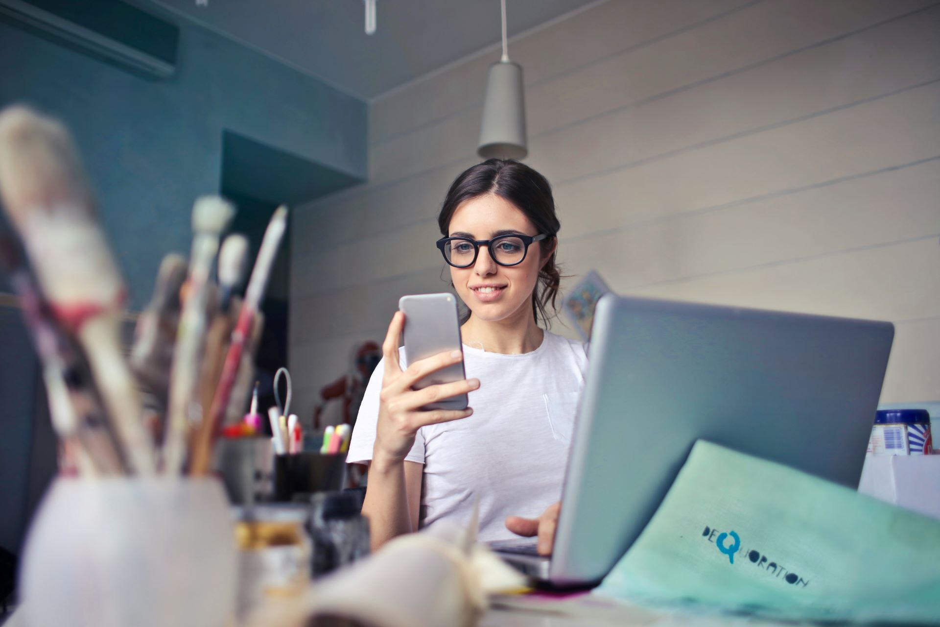 Eine junge Frau mit Brille arbeitet an einem Laptop und schaut gleichzeitig auf ihr Smartphone, umgeben von Bastelmaterialien auf einem Schreibtisch.