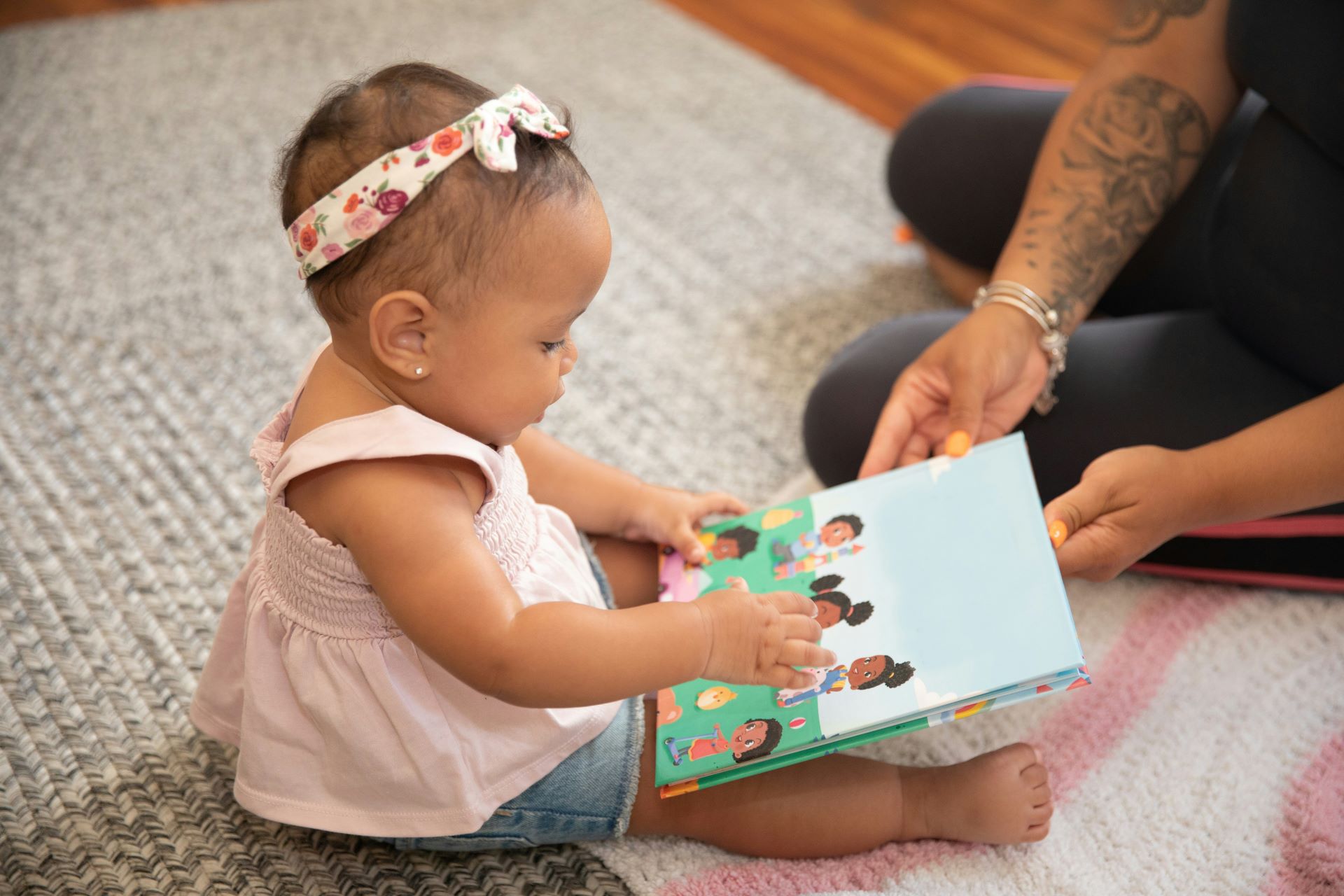 Ein Baby sitzt auf einem Teppich und schaut sich ein farbenfrohes Kinderbuch an, das von einer erwachsenen Person gehalten wird.