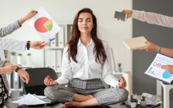 Frau sitzt im Buero im Schneidersitz auf dem Tisch und meditiert, waehrend Kolleginnen Unterlagen, Handy und Diagramme in der Hand halten – Symbol fuer Stressbewaeltigung am Arbeitsplatz