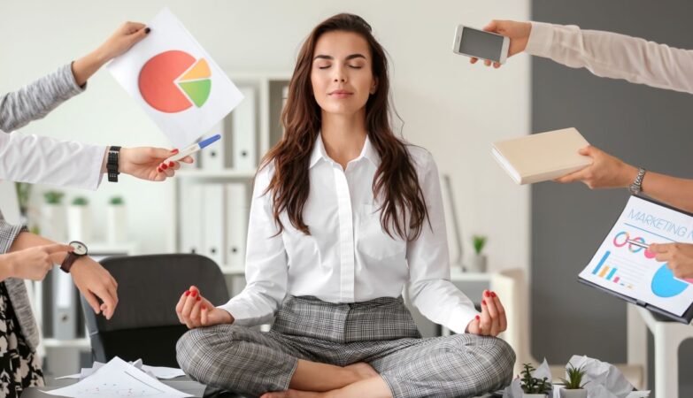 Frau sitzt im Buero im Schneidersitz auf dem Tisch und meditiert, waehrend Kolleginnen Unterlagen, Handy und Diagramme in der Hand halten – Symbol fuer Stressbewaeltigung am Arbeitsplatz
